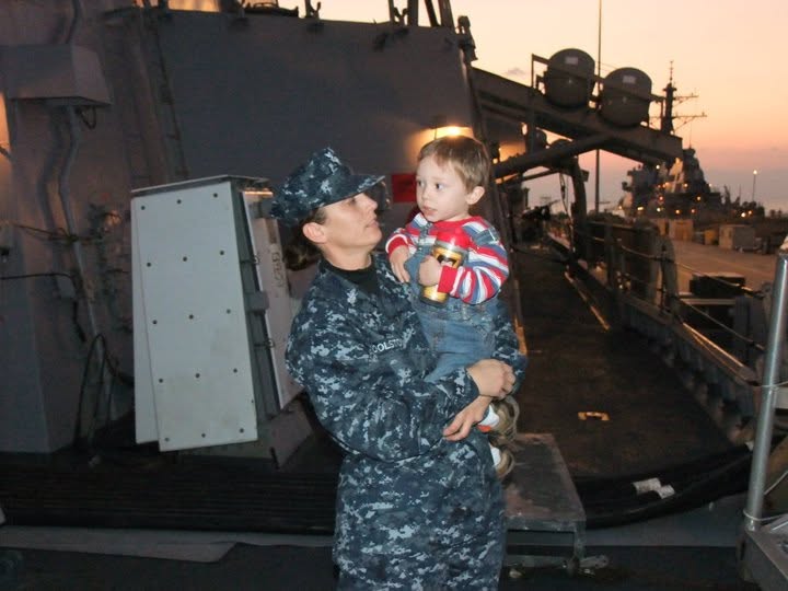 Ian as a toddler held by his mom, in Navy working uniform, aboard a ship at sunset